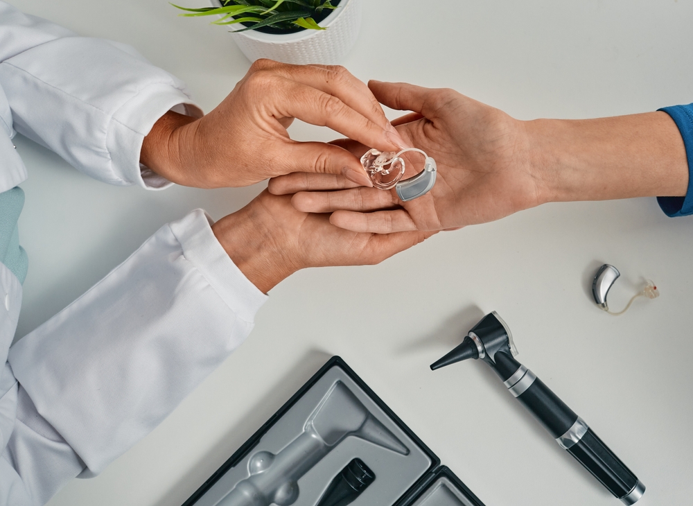 A healthcare professional hands a hearing aid to a patient. Medical tools and another hearing aid are on the white table, with a small plant in the background.