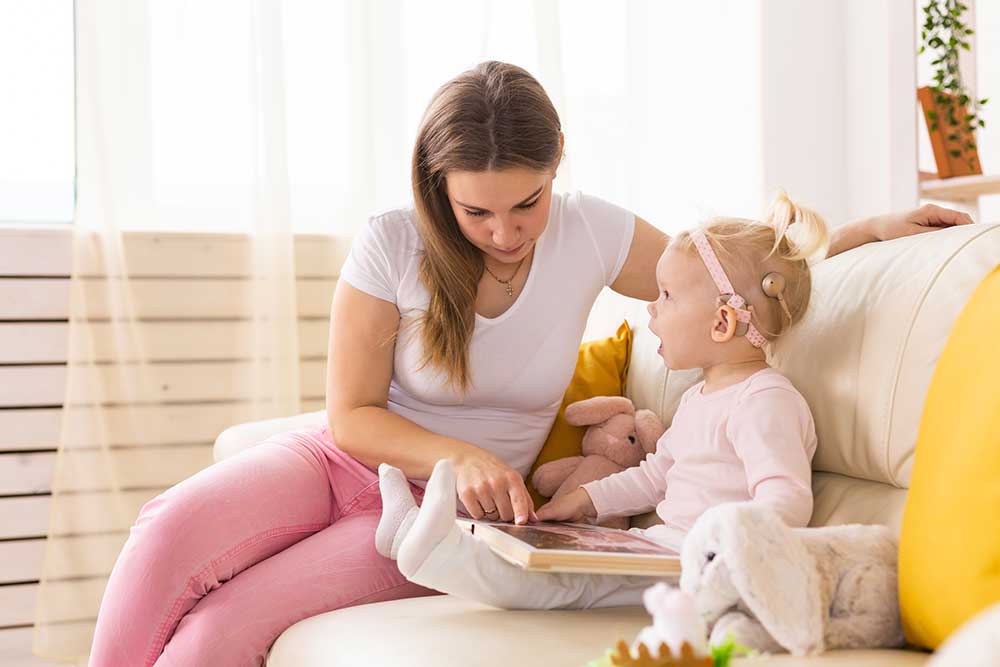 A woman and a young girl with a hearing aid sit on a couch reading a book together. The child holds a stuffed animal, and both appear engaged in the story. The setting is bright and cozy.