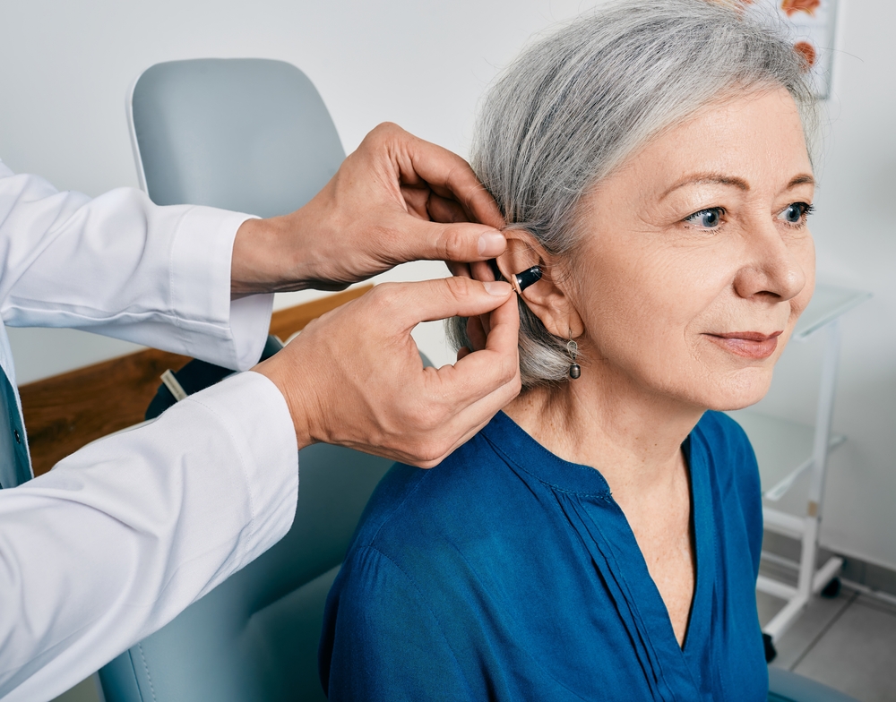 A healthcare professional fits a hearing aid onto the ear of an older woman with gray hair, who is sitting in a medical office and smiling slightly.