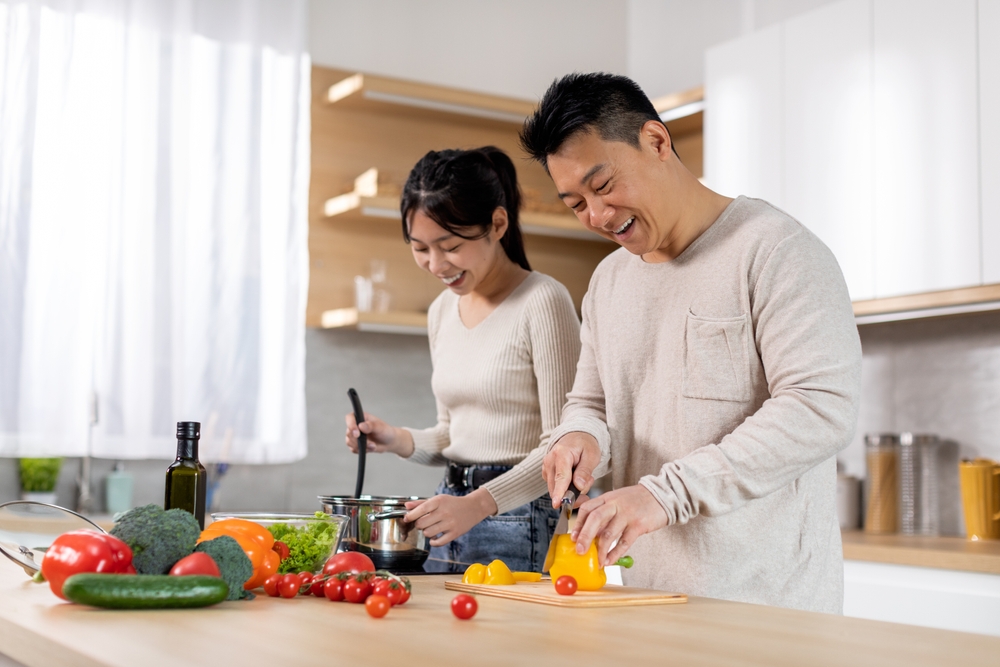 A man slices a yellow bell pepper while a woman stirs food in a pot. Both are smiling and preparing vegetables together in a bright, modern kitchen. Fresh produce is arranged on the counter.