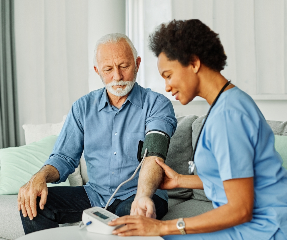 A nurse in blue scrubs checks the blood pressure of an older man with gray hair and mustache, who is sitting on a couch at home.