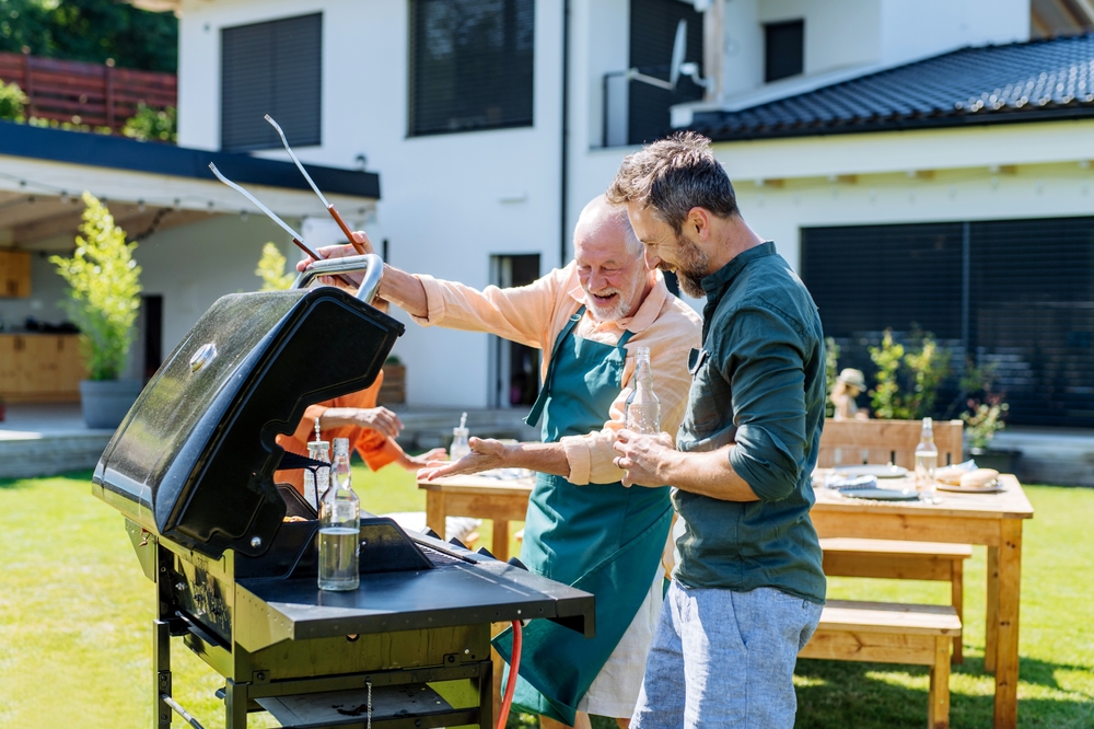 Two men stand beside a grill in a backyard, smiling and cooking together. One wears an apron and holds tongs, while the other looks at the food. A picnic table and a modern house are in the background.