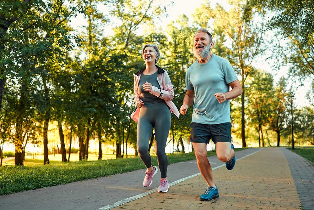 A smiling older man and woman jog side by side on a paved path in a park, surrounded by green trees and sunlight. Both wear athletic clothing and look happy and energetic.