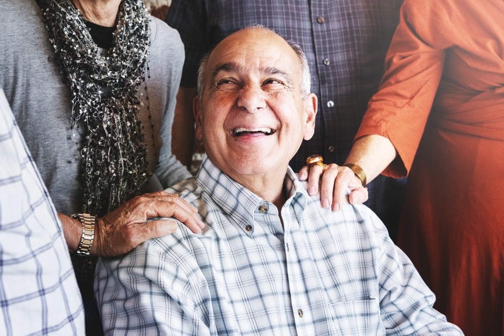 An older man wearing a plaid shirt smiles joyfully as several people stand around him, resting their hands on his shoulders in a gesture of support and affection.