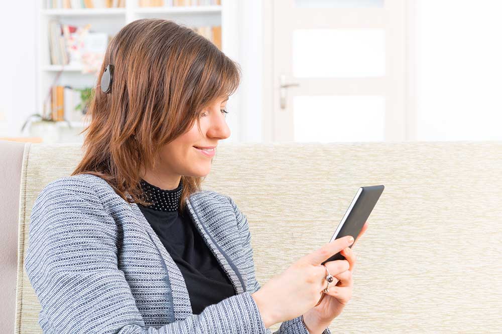 A woman with a cochlear implant sits on a couch, smiling while using a smartphone or tablet. Bookshelves and a bright doorway are visible in the background.