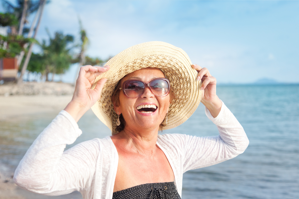 Smiling woman wearing sunglasses and a sunhat stands on a sunny beach with palm trees and the ocean in the background, enjoying the pleasant weather.