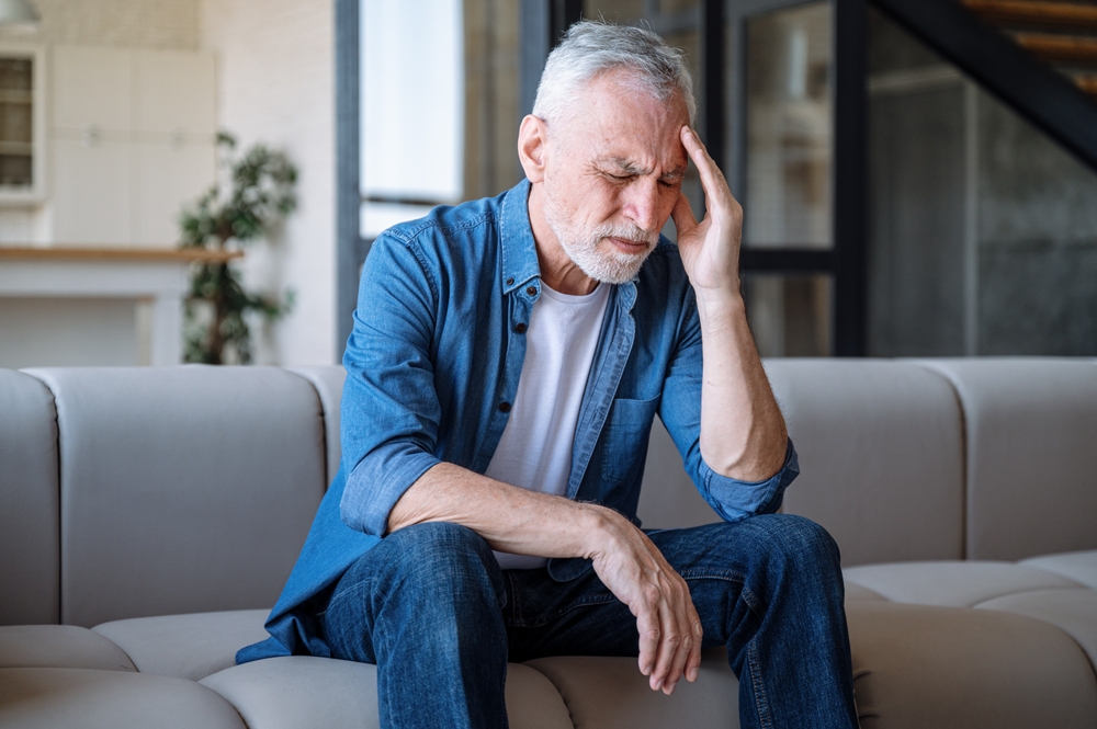 An older man with gray hair sits on a couch, eyes closed, touching his forehead with a pained expression, suggesting he may have a headache or be feeling unwell.