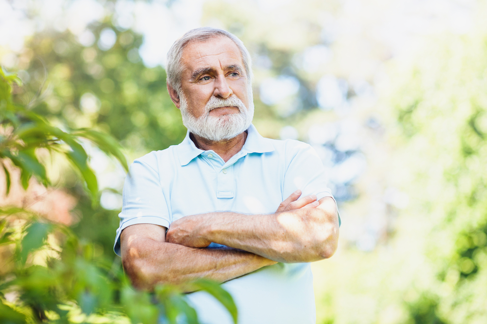 An older man with a white beard and mustache stands outdoors with his arms crossed, wearing a light blue polo shirt. He looks thoughtful, surrounded by greenery and soft sunlight.