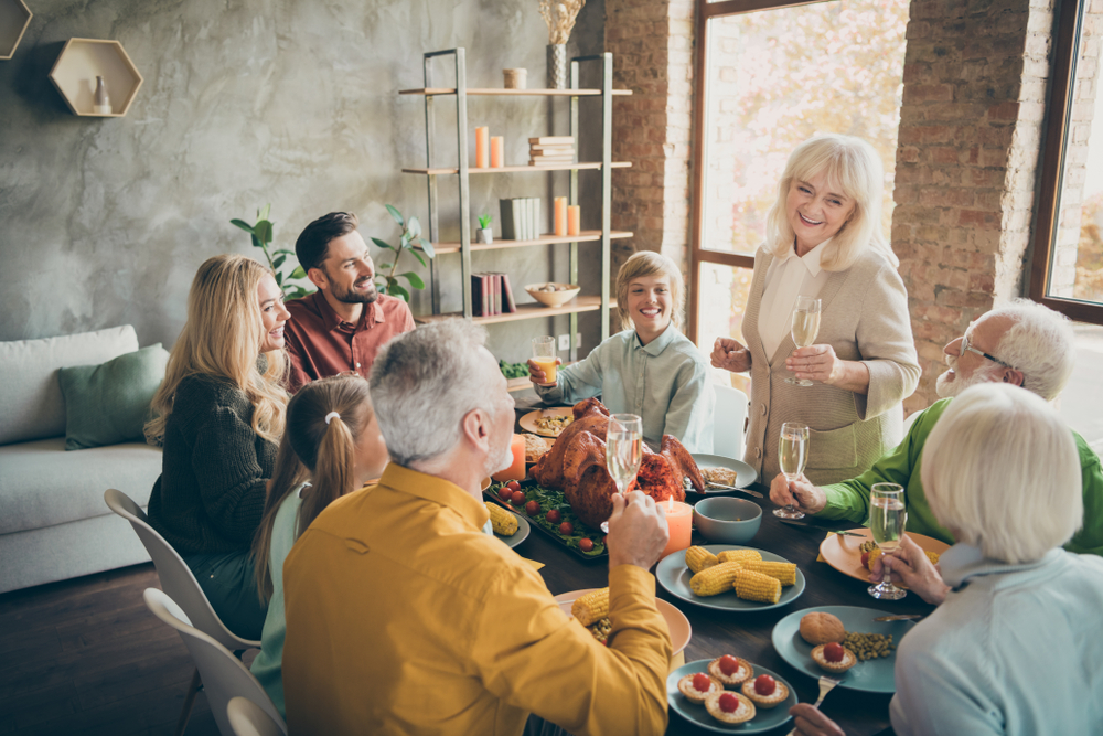 A group of people, including children and older adults, sit around a festive dinner table with food and drinks, smiling and toasting together in a warmly lit room.