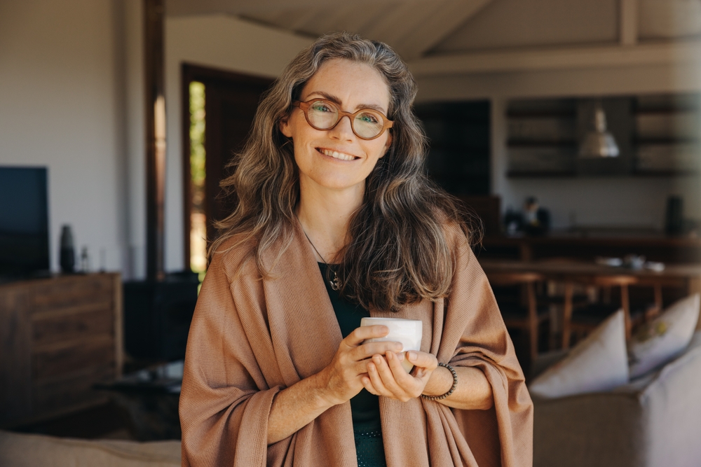 A smiling woman with long gray hair and glasses stands indoors, wearing a light brown shawl and holding a white mug in both hands. The background shows a cozy, modern living space.