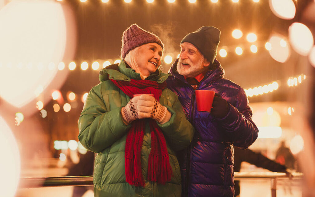 An elderly couple dressed in winter coats, hats, and gloves smile at each other under festive lights at night. The man holds a red mug, and the background is blurred with warm, glowing bokeh.