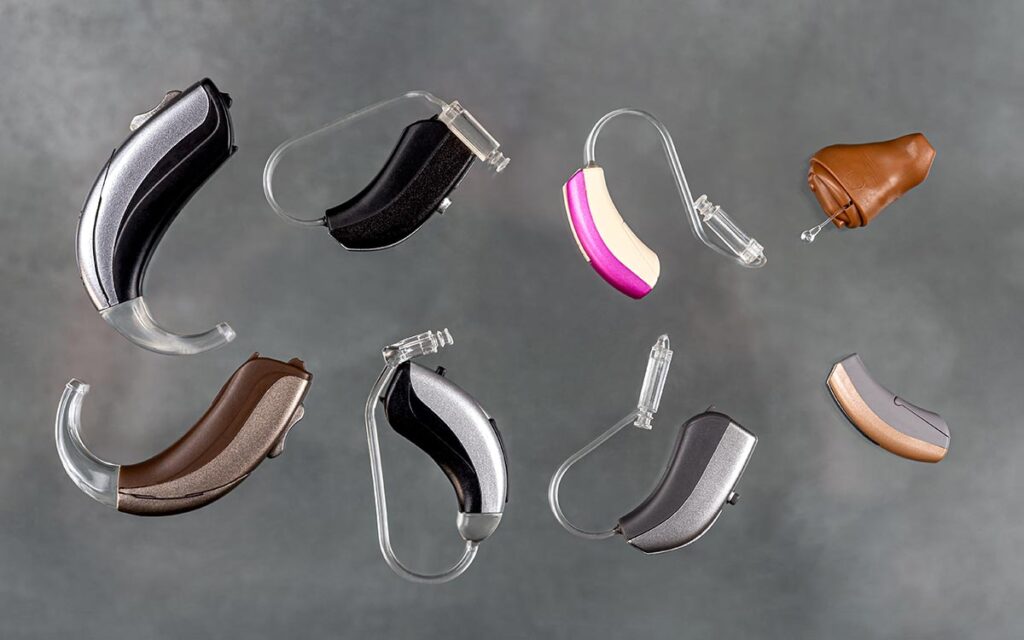 A group of eight hearing aids in various shapes, sizes, and colors arranged on a gray background. Some have flesh-toned, black, silver, or pink accents, and feature clear plastic ear tubes or molds.