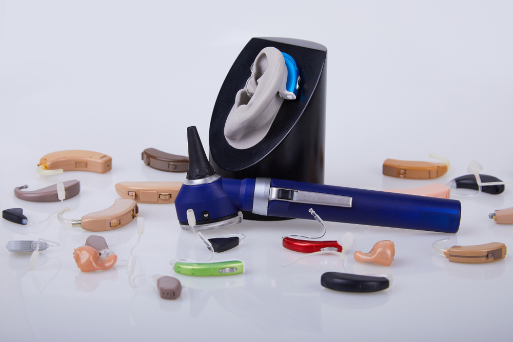 Various hearing aids in different colors are scattered on a white surface, with an ear model wearing a blue hearing aid and an otoscope placed in front.
