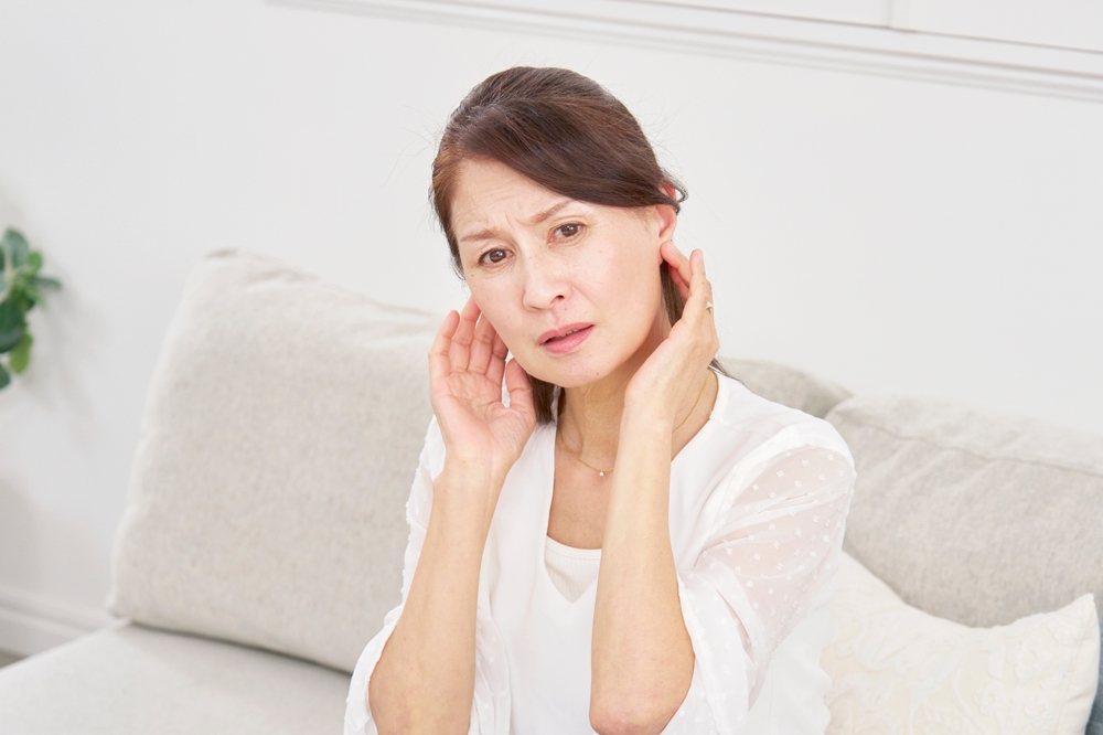 A middle-aged woman sitting on a couch touching her ears with both hands and looking concerned, as if experiencing ear discomfort or pain.